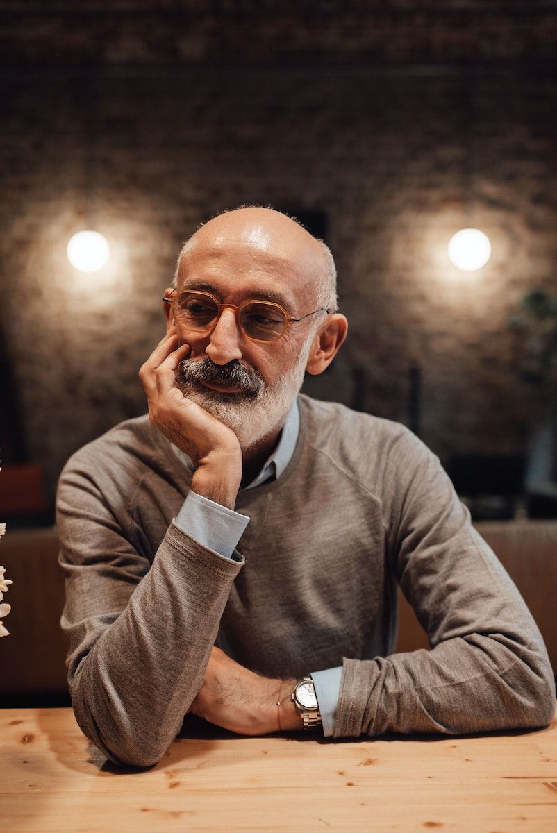 Pensive bald elderly male with gray beard and eyeglasses looking away while sitting at wooden table and leaning on hand